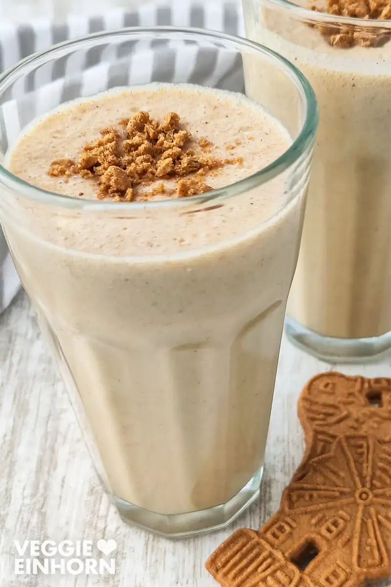 Veganer Spekulatius Milchshake Nahaufnahme eines veganen Spekulatius-Milchshakes. Daneben liegt ein Spekulatius-Keks auf einem hellen Holztisch und im Hintergrund steht ein zweites Glas mit Milchshake.