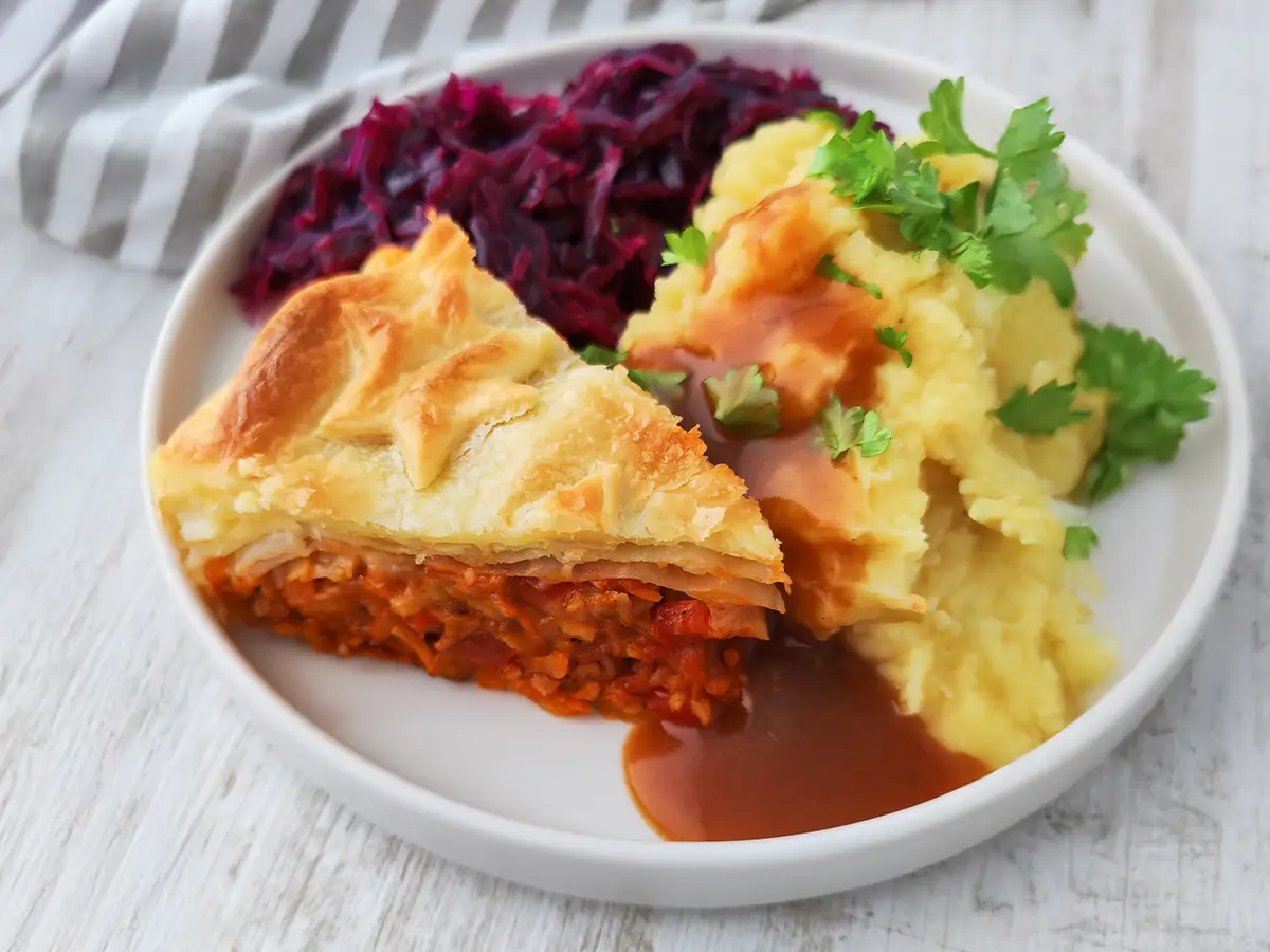 Ein Teller mit einem Stück veganem Blätterteigbraten, Kartoffelpüree mit brauner Soße und Rotkohl.