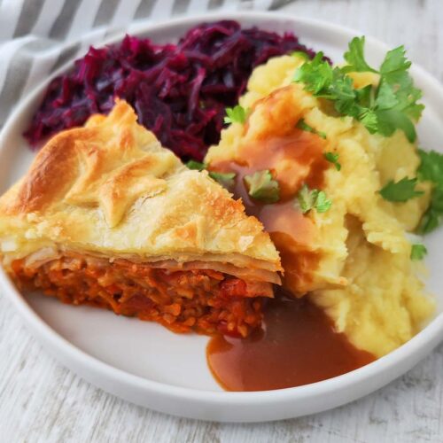 Ein Stück veganer Blätterteigbraten liegt auf einem Teller zusammen mit Kartoffelpüree, brauner Soße und Rotkohl.