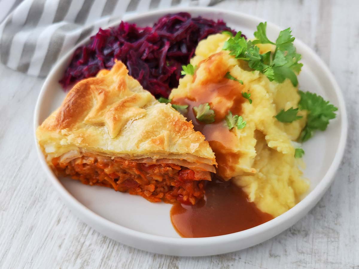 Ein Stück veganer Blätterteigbraten liegt auf einem Teller zusammen mit Kartoffelpüree, brauner Soße und Rotkohl.