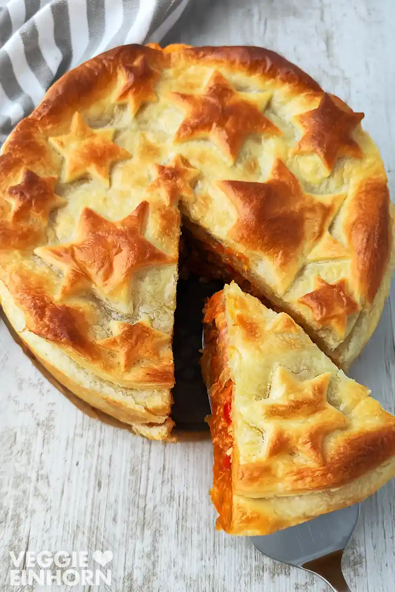 Ein veganer Tofu Hackbraten mit goldbraunem Blätterteig, verziert mit ausgestochenen Sternen. Ein Stück wird heraus gehoben.