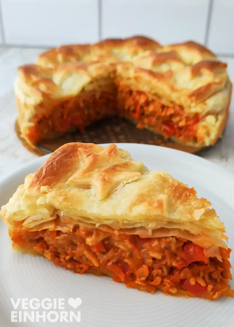 Ein Stück veganer Blätterteigbraten liegt auf einem Teller, während der restliche Braten im Hintergrund angeschnitten zu sehen ist.