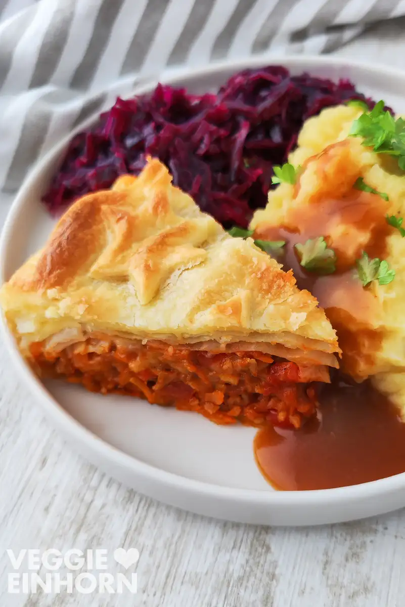 Ein Stück veganer Hackbraten mit Räuchertofu, Blätterteig und Wirsing ist serviert auf einem festlichen Teller mit Rotkohl, Kartoffelpüree und veganer Bratensoße.