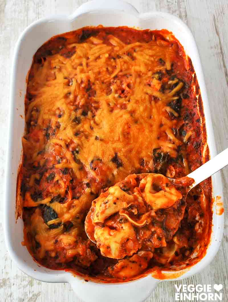 Eine weiße Auflaufform mit veganem Tortellini-Auflauf mit Spinat, Tomatensoße und veganem Käse.