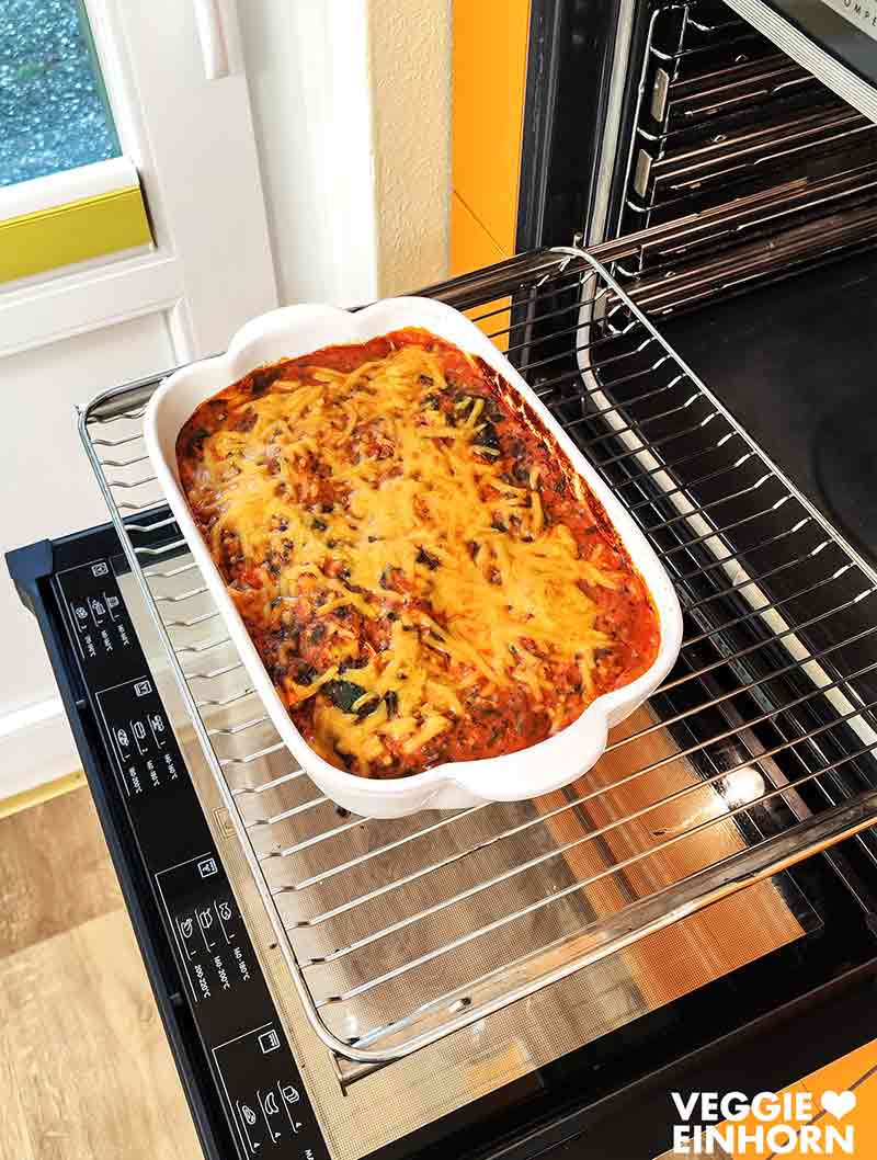Ein veganer Tortellini-Auflauf mit Spinat steht fertig gebacken auf dem Rost eines geöffneten Backofens, goldgelb überbacken.