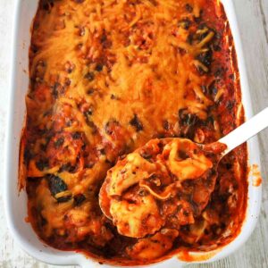 Veganer Tortellini-Auflauf mit Spinat in einer weißen Auflaufform, bedeckt mit veganem Käse, dazu ein Löffel, der eine Portion Tortellini entnimmt.