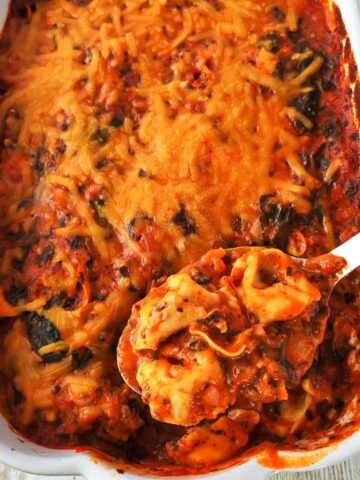 Veganer Tortellini-Auflauf mit Spinat in einer weißen Auflaufform, bedeckt mit veganem Käse, dazu ein Löffel, der eine Portion Tortellini entnimmt.