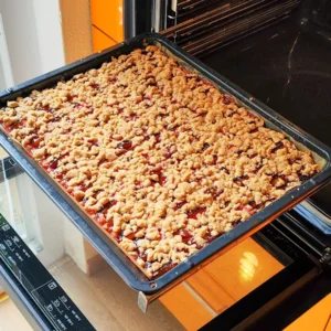 Ein großes Backblech mit veganem Zwetschgenkuchen, der mit goldbraunen Zimtstreuseln bedeckt ist und im geöffneten Backofen steht.