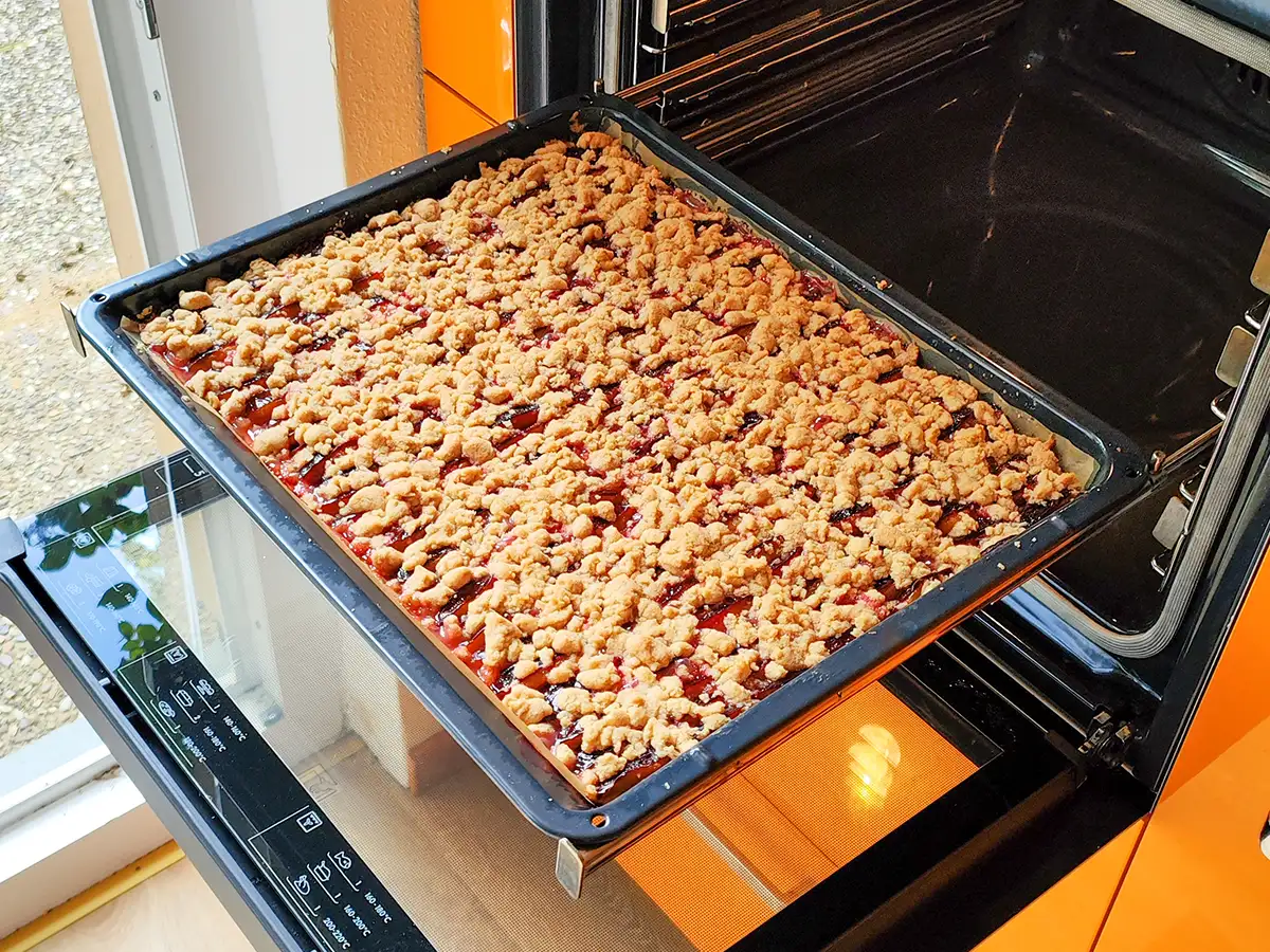 Ein großes Backblech mit veganem Zwetschgenkuchen, der mit goldbraunen Zimtstreuseln bedeckt ist und im geöffneten Backofen steht.
