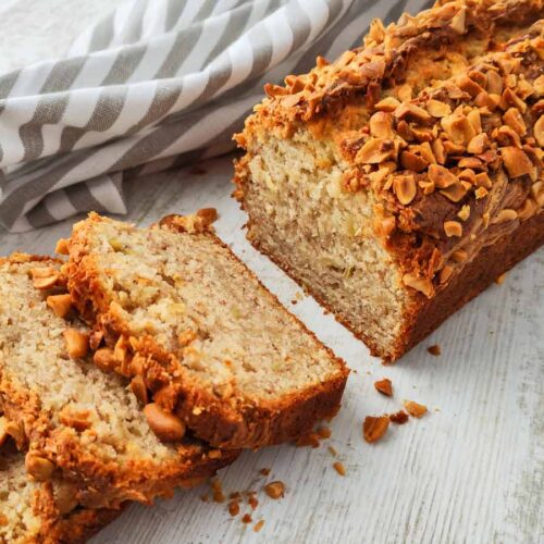 Ein veganes Bananenbrot mit Erdnüssen ist teilweise in Scheiben geschnitten und liegt auf einer hellen Holzoberfläche mit einem gestreiften Tuch im Hintergrund.