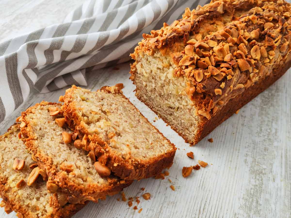 Ein veganes Bananenbrot mit Erdnüssen ist teilweise in Scheiben geschnitten und liegt auf einer hellen Holzoberfläche mit einem gestreiften Tuch im Hintergrund.