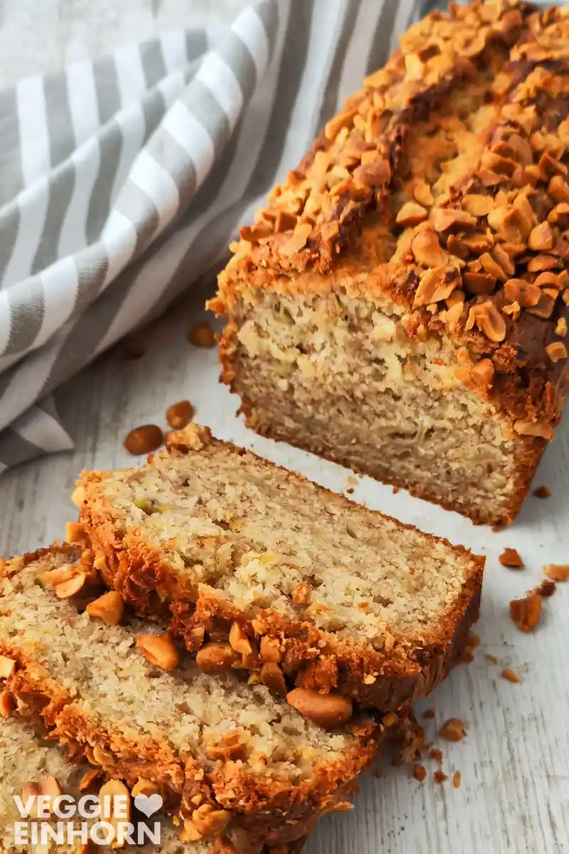 Angeschnittenes veganes Bananenbrot mit Erdnussbutter und gehackten Erdnüssen auf einer hellen Holzoberfläche, daneben ein weiß-grau gestreiftes Tuch.