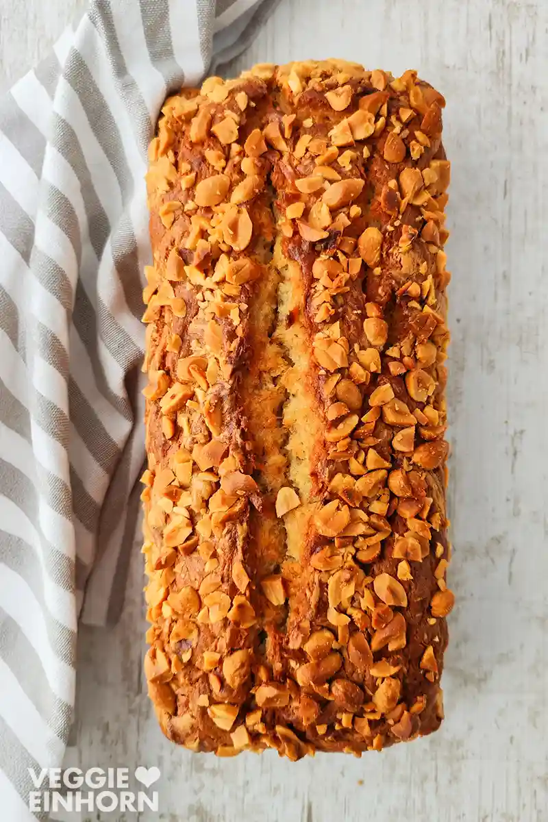 Ein ganzes veganes Bananenbrot mit Erdnussstückchen liegt auf einer hellen Holzfläche, daneben ein gestreiftes Tuch.