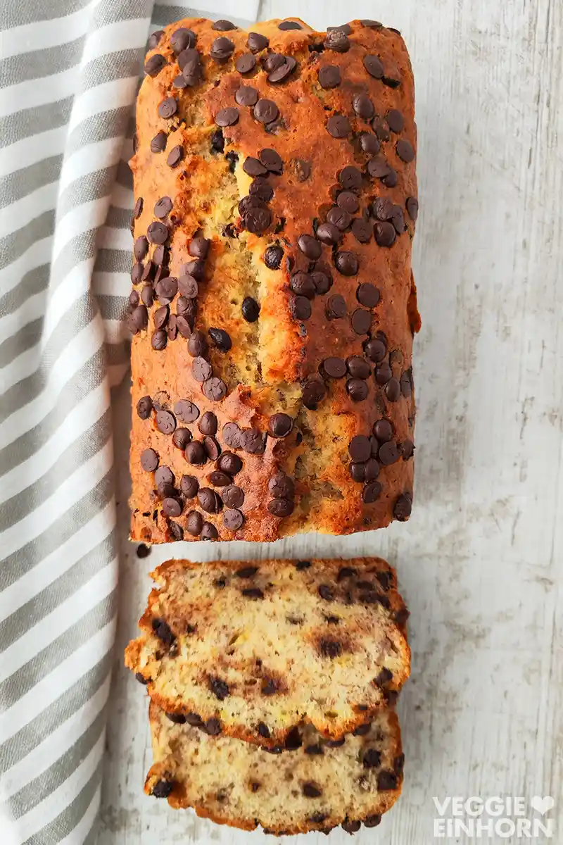 Veganes Bananenbrot mit Schokotropfen von oben fotografiert, daneben ein gestreiftes Tuch auf hellem Holz.