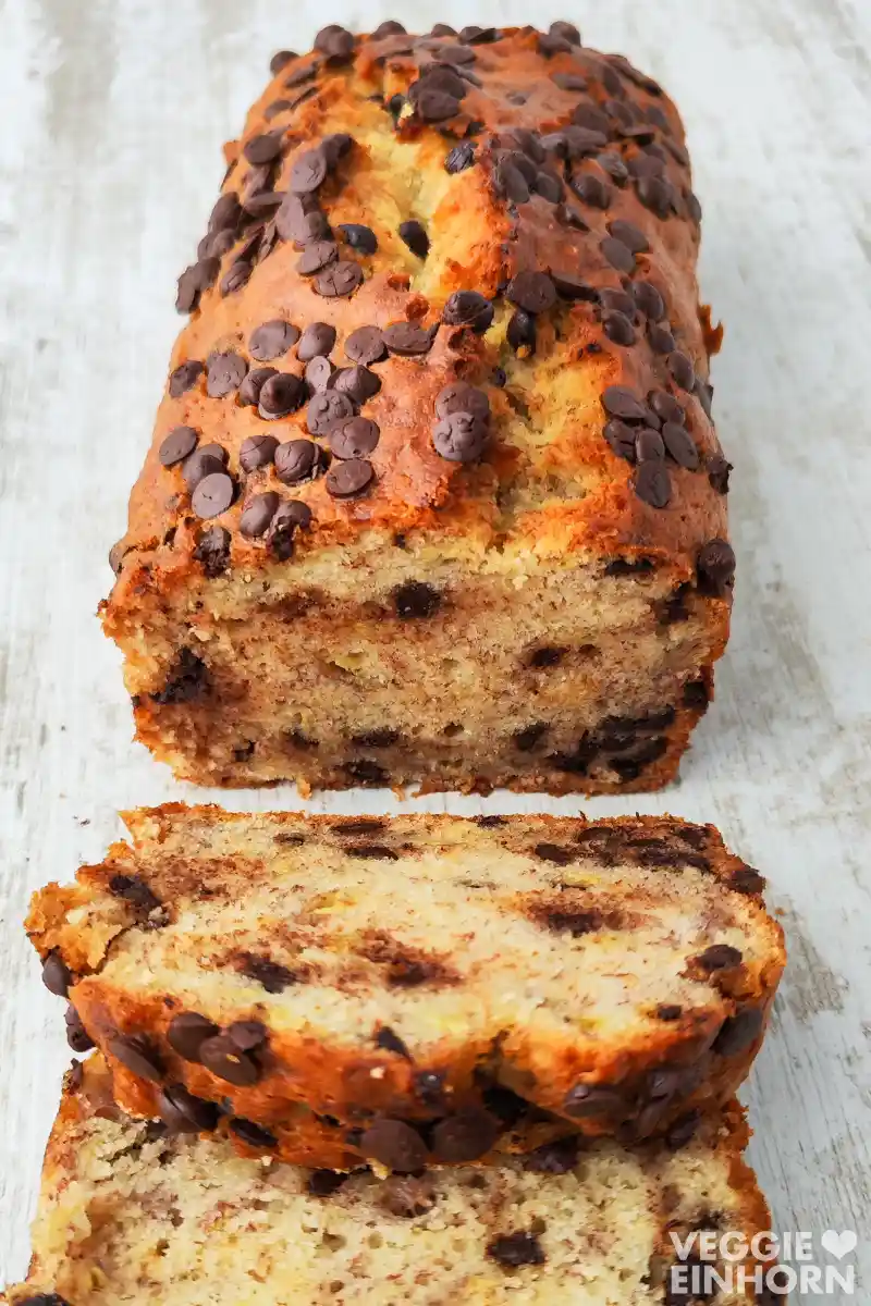 Ein veganes Bananenbrot mit vielen Schokotropfen liegt angeschnitten auf einem hellen Holztisch.