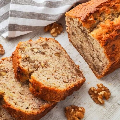 Ein veganes Bananenbrot mit Walnüssen liegt angeschnitten auf einer hellen Holzoberfläche, daneben sind einzelne Walnusskerne verteilt.