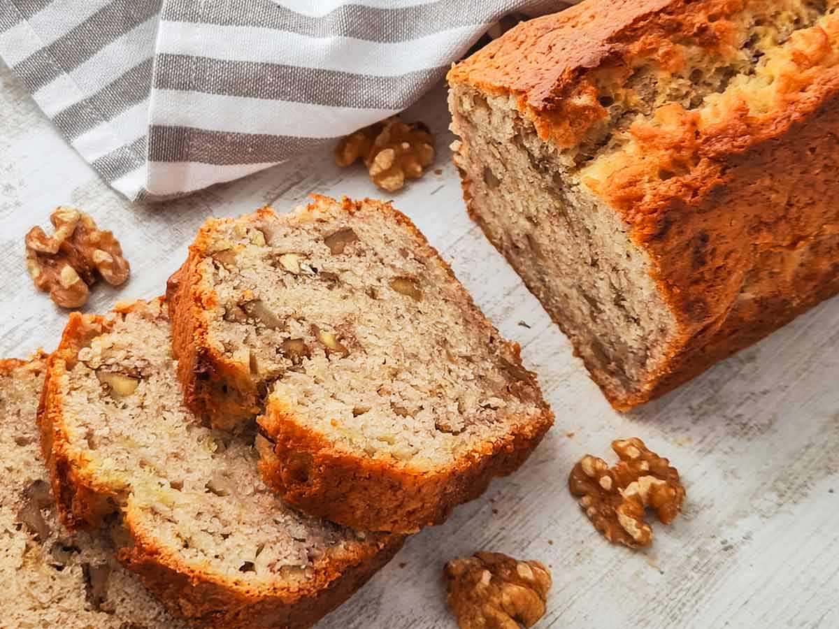 Ein veganes Bananenbrot mit Walnüssen liegt angeschnitten auf einer hellen Holzoberfläche, daneben sind einzelne Walnusskerne verteilt.