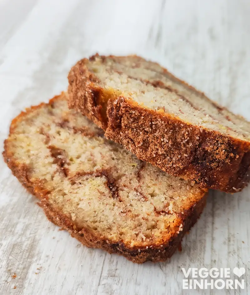 Zwei Scheiben veganes Bananenbrot mit Zimt liegen auf einem weißen Holztisch.