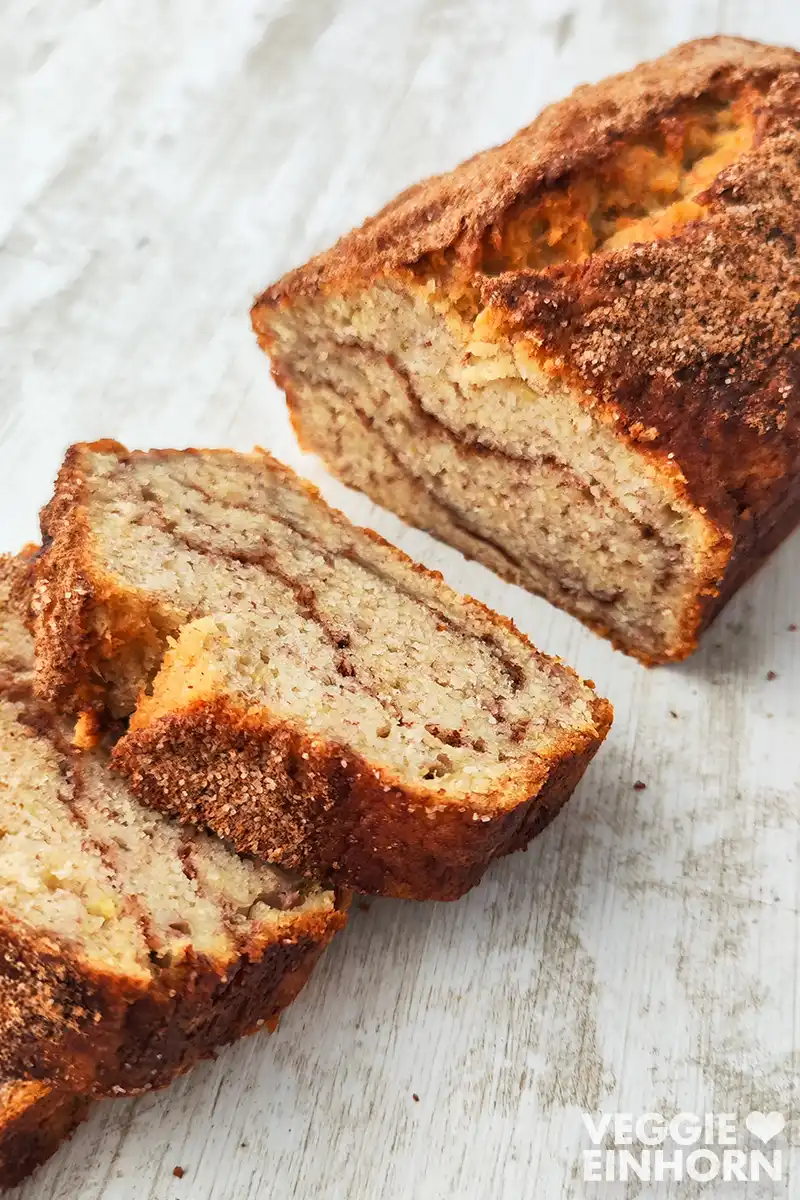 Ein veganes Bananenbrot mit Zimt liegt angeschnitten auf einer weißen Holzoberfläche.