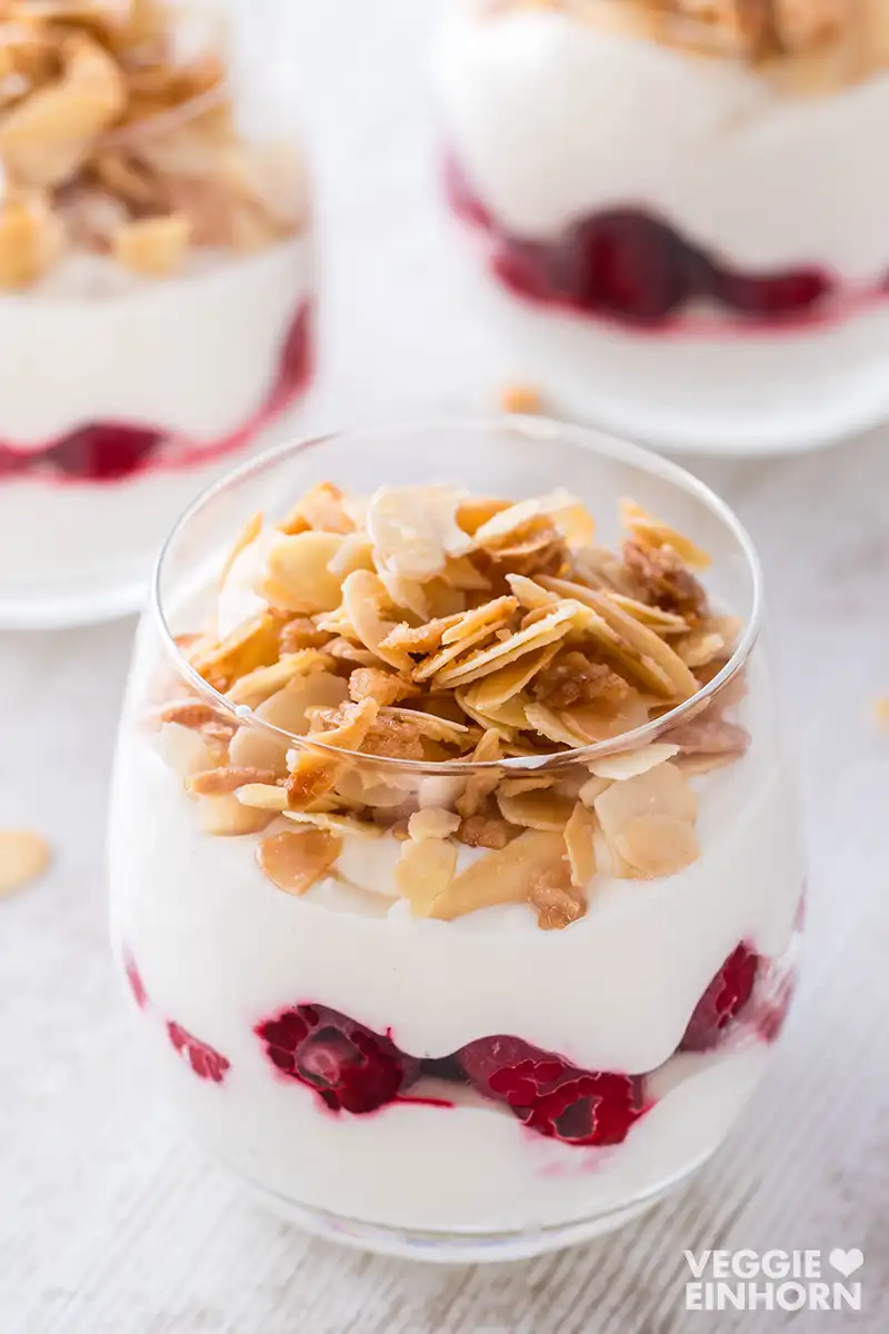 Ein Glas mit veganem Himbeer-Dessert, geschichtet mit Dessertcreme und Himbeeren, getoppt mit karamellisierten Mandelblättchen, im Hintergrund weitere Dessertgläser.