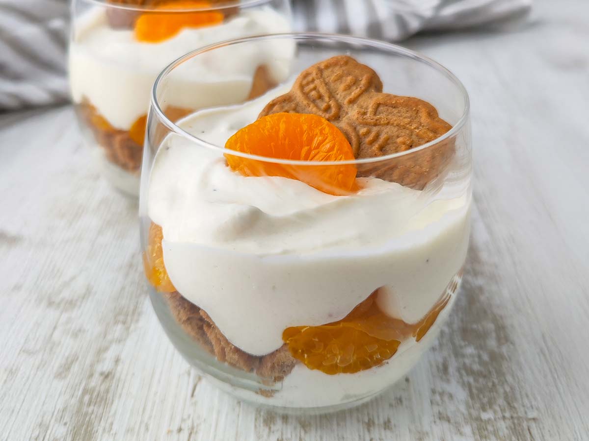 Ein veganes Schichtdessert im Glas mit Dessertcreme, Mandarinen und Spekulatius, fotografiert auf hellem Untergrund.