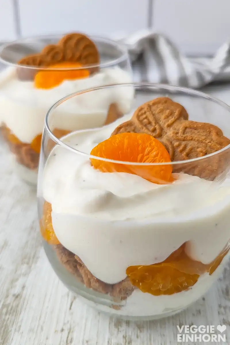 Zwei Gläser mit veganem Weihnachtsdessert aus Spekulatiuskeksen, Mandarinen und Dessertcreme auf einem weißen Tisch.
