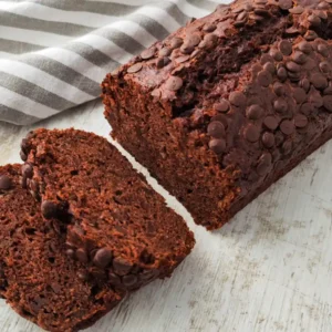 Ein saftiges veganes Schoko-Bananenbrot mit vielen Schokotropfen liegt angeschnitten auf einer hellen Holzfläche, daneben ein grau gestreiftes Tuch.