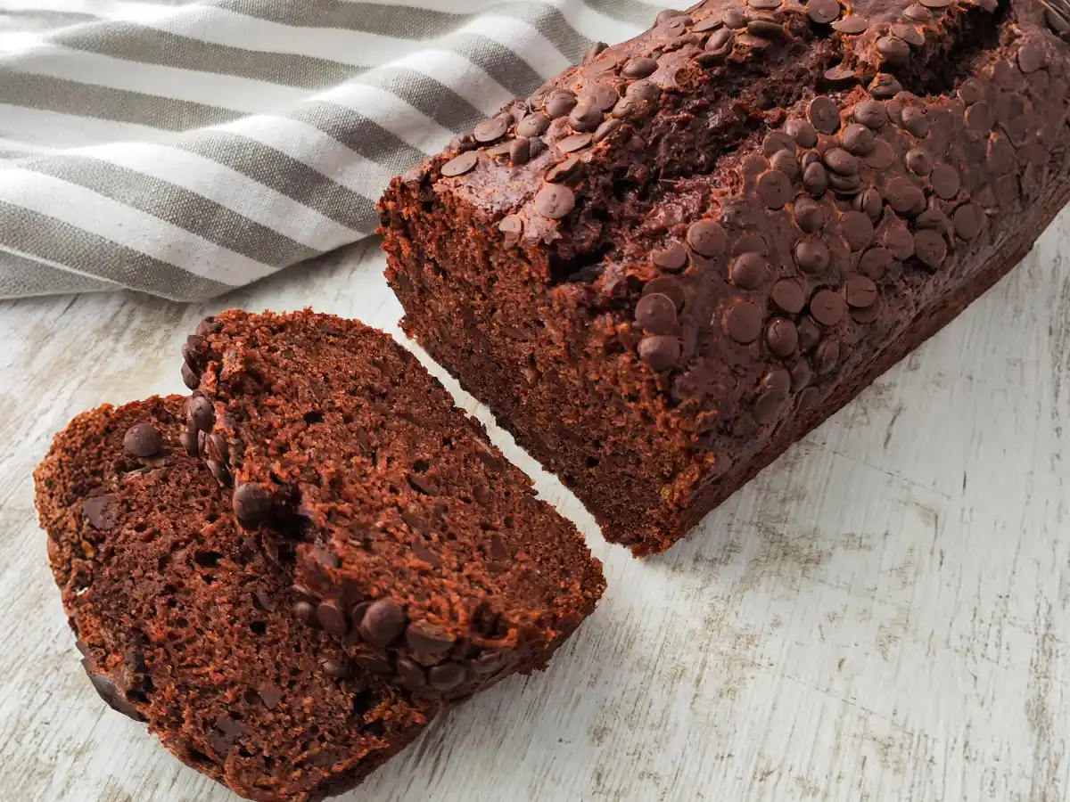 Ein saftiges veganes Schoko-Bananenbrot mit vielen Schokotropfen liegt angeschnitten auf einer hellen Holzfläche, daneben ein grau gestreiftes Tuch.