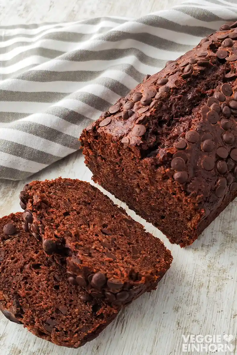 Veganes Schoko Bananenbrot Ein saftiges veganes Schoko-Bananenbrot mit Schokotropfen liegt auf einer hellen Holzoberfläche, daneben ein gestreiftes Küchentuch.