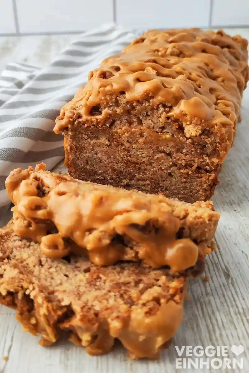 Veganes Spekulatius Bananenbrot mit cremiger Karamellglasur, aufgeschnitten auf einem hellen Holztisch neben einem gestreiften Küchentuch.