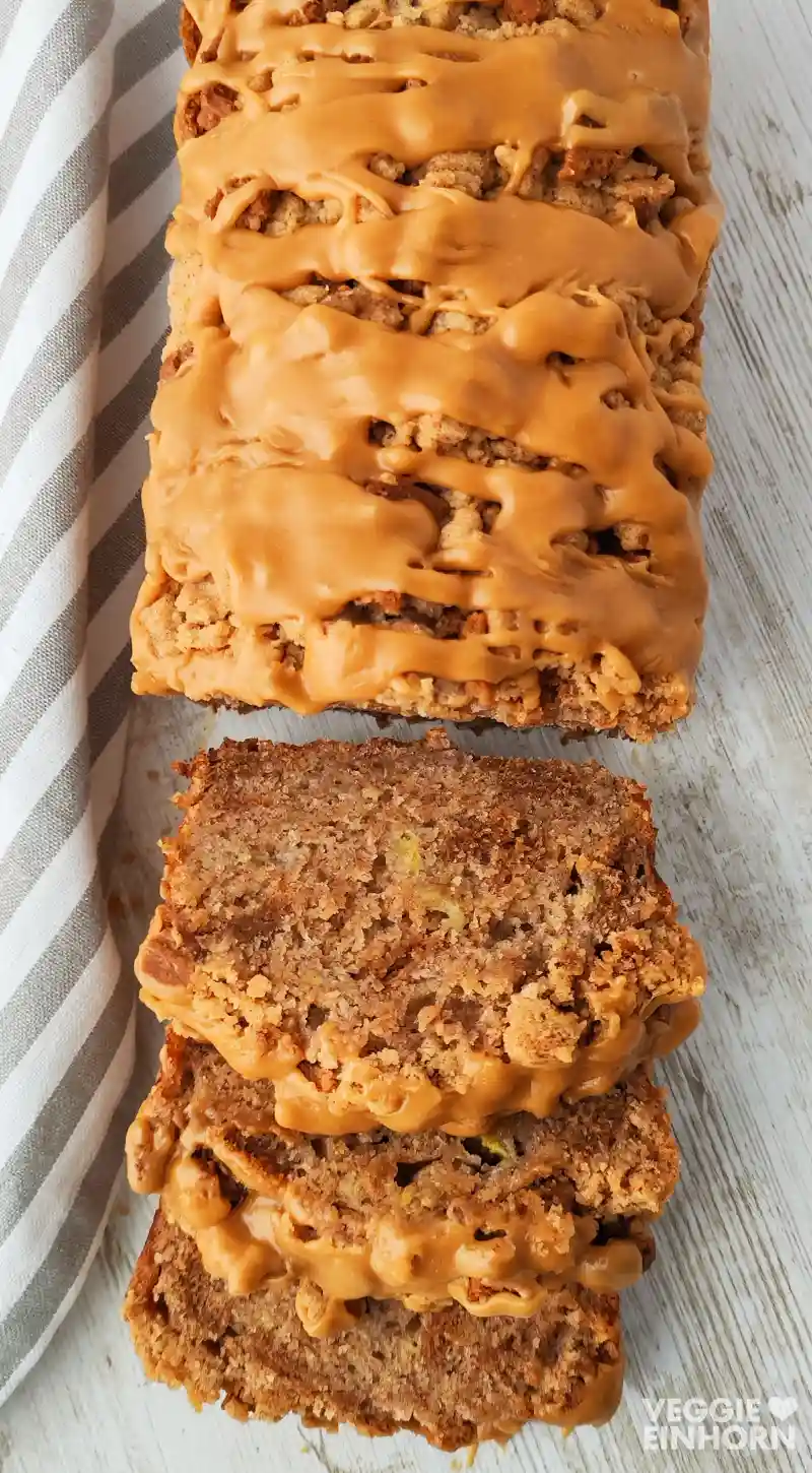 Veganes Spekulatius Bananenbrot mit dicker Karamellglasur, angeschnitten auf einem weißen Holztisch mit grau-weiß gestreiftem Tuch.