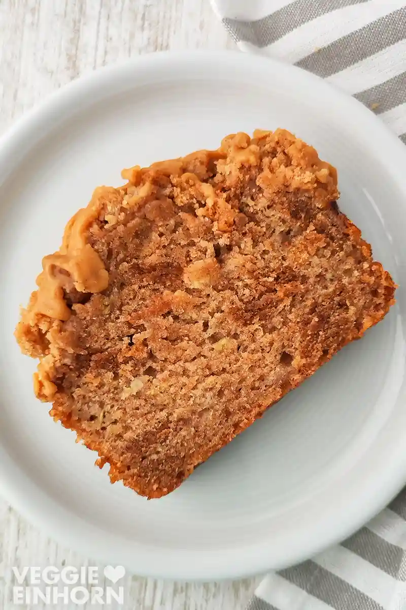 Ein Stück veganes Spekulatius Bananenbrot mit Karamellglasur liegt auf einem weißen Teller, daneben ein gestreiftes Tuch auf hellem Holz.