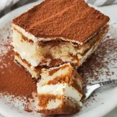 Ein Stück veganes Tiramisu mit dicker Kakaoschicht auf der Oberfläche liegt auf einem weißen Teller.