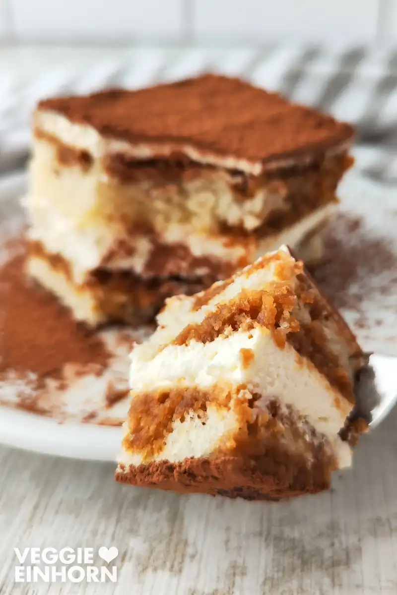 Ein Stück veganes Tiramisu liegt auf einem weißen Teller, im Vordergrund befindet sich ein kleines Stück des Tiramisus auf einer Gabel.