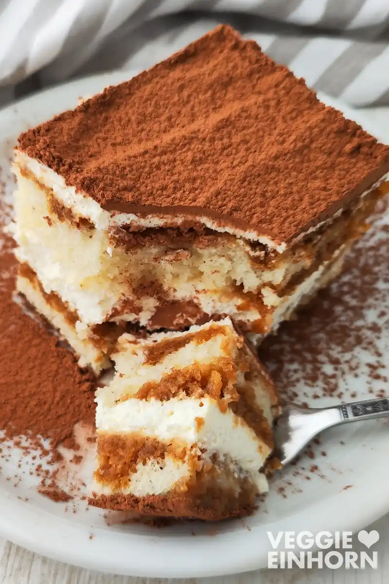 Veganes Tiramisu mit veganem Frischkäse Ein Stück veganes Tiramisu liegt auf einem weißen Teller, im Vordergrund ein Stück des Tiramisus auf einer Gabel.