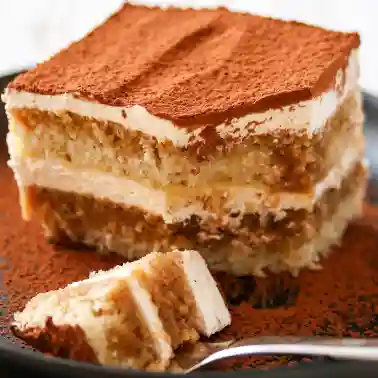 Nahaufnahme eines Stücks veganen Tiramisus mit Schichten aus Creme und in Kaffee getränktem Biskuit, bestäubt mit Kakaopulver, auf einem Teller serviert, vorne ein kleines Stück auf einer Gabel.