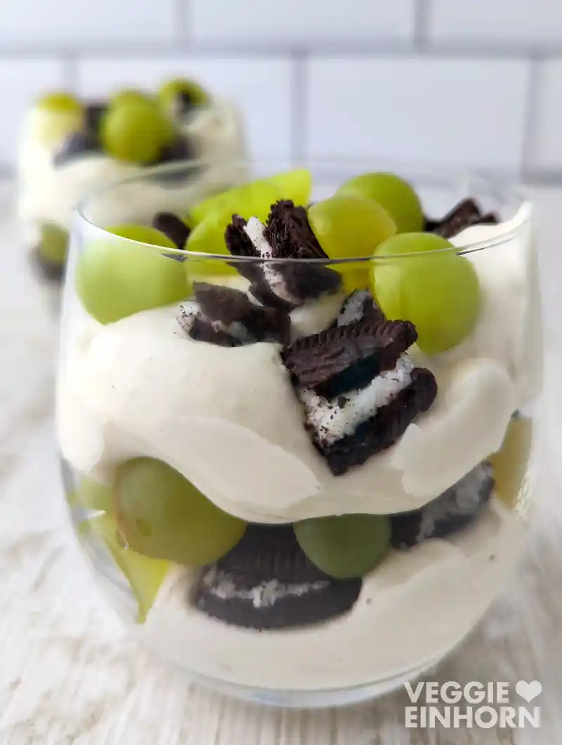 Ein Glas mit cremigem veganem Dessert, geschichtet mit grünen Weintrauben und zerbrochenen Oreo-Keksen auf hellem Untergrund.