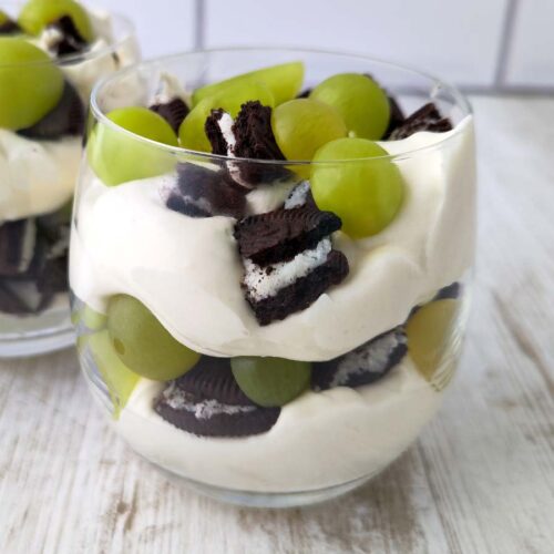 Nahaufnahme eines Glases mit veganem Dessert aus Dessertcreme, grünen Weintrauben und Oreo-Stücken, im Hintergrund ein weiteres Glas.