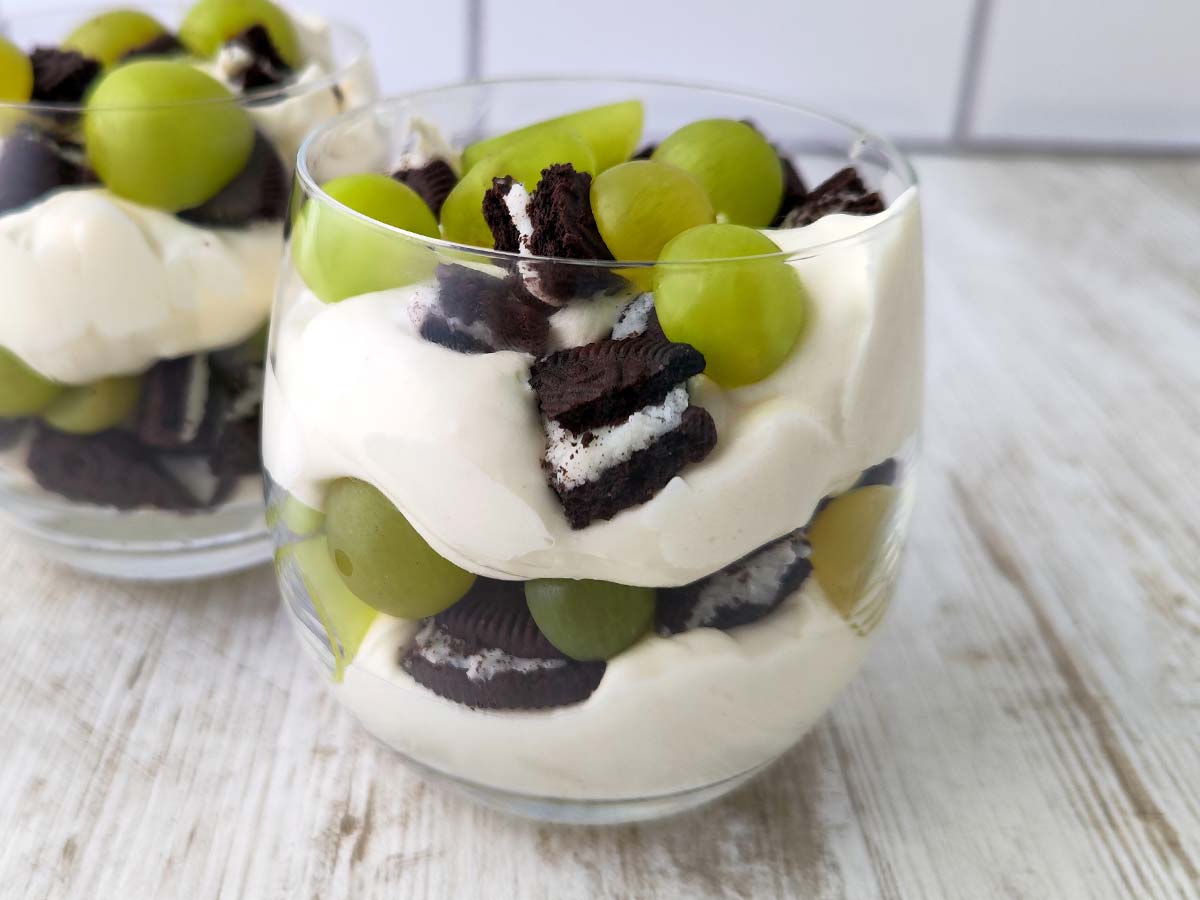 Nahaufnahme eines Glases mit veganem Dessert aus Dessertcreme, grünen Weintrauben und Oreo-Stücken, im Hintergrund ein weiteres Glas.