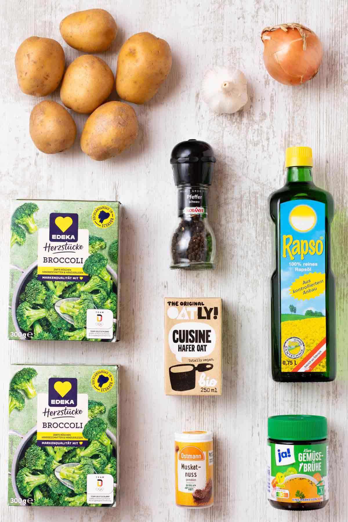 Zutaten für veganen Kartoffel-Brokkoli-Auflauf auf einem Holztisch – Kartoffeln, Brokkoli, vegane Hafer-Sahne, Zwiebel, Knoblauch, Muskat, Gemüsebrühe, Rapsöl und Pfeffer nebeneinander angeordnet.