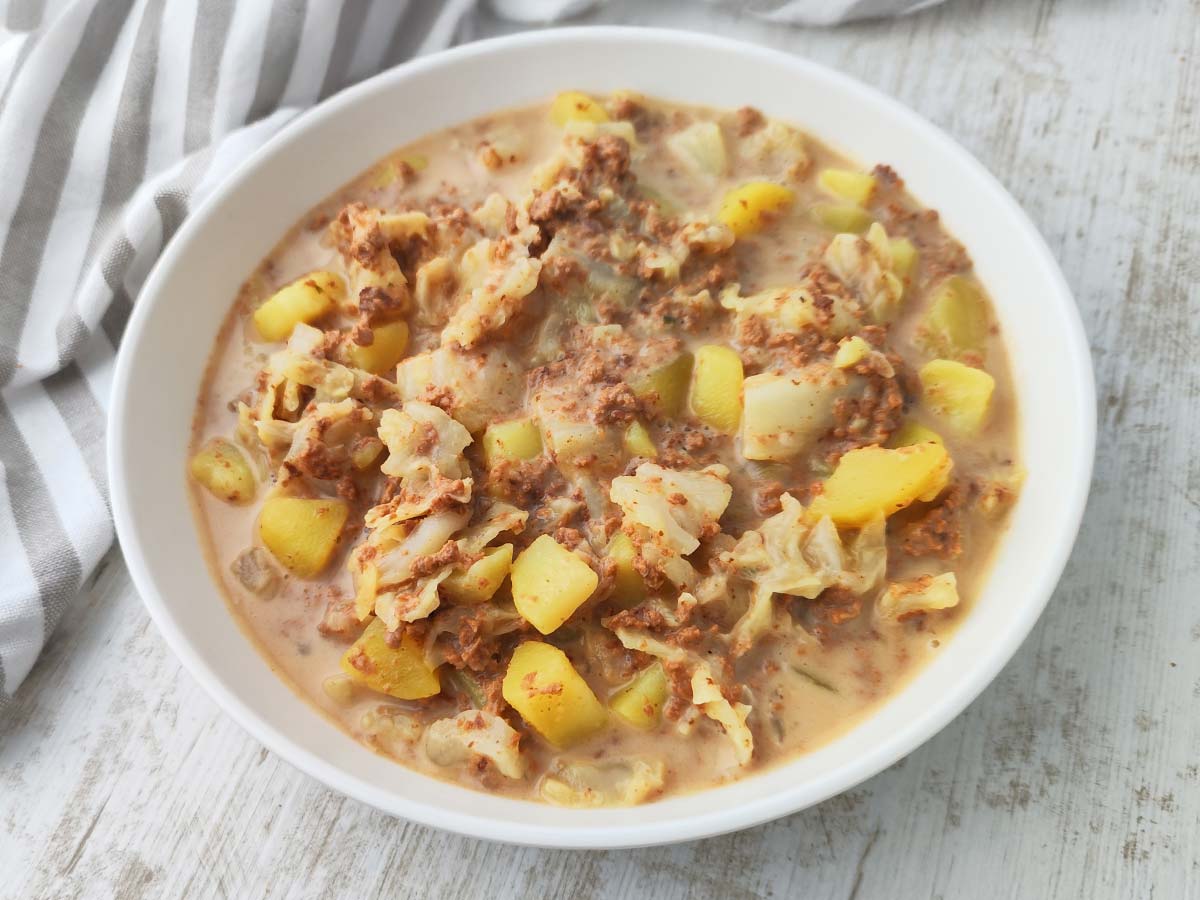 Ein Teller mit veganem Wirsingeintopf mit Kartoffeln und veganem Hack steht auf einem hellen Holztisch.