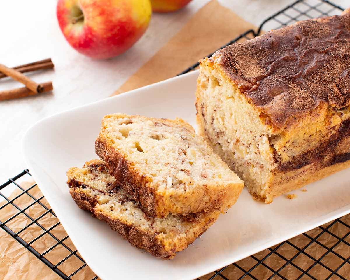 Ein veganer Zimtkuchen mit Apfel, angeschnitten auf einer weißen Kuchenplatte. Im Hintergrund liegen frische Äpfel und Zimtstangen.