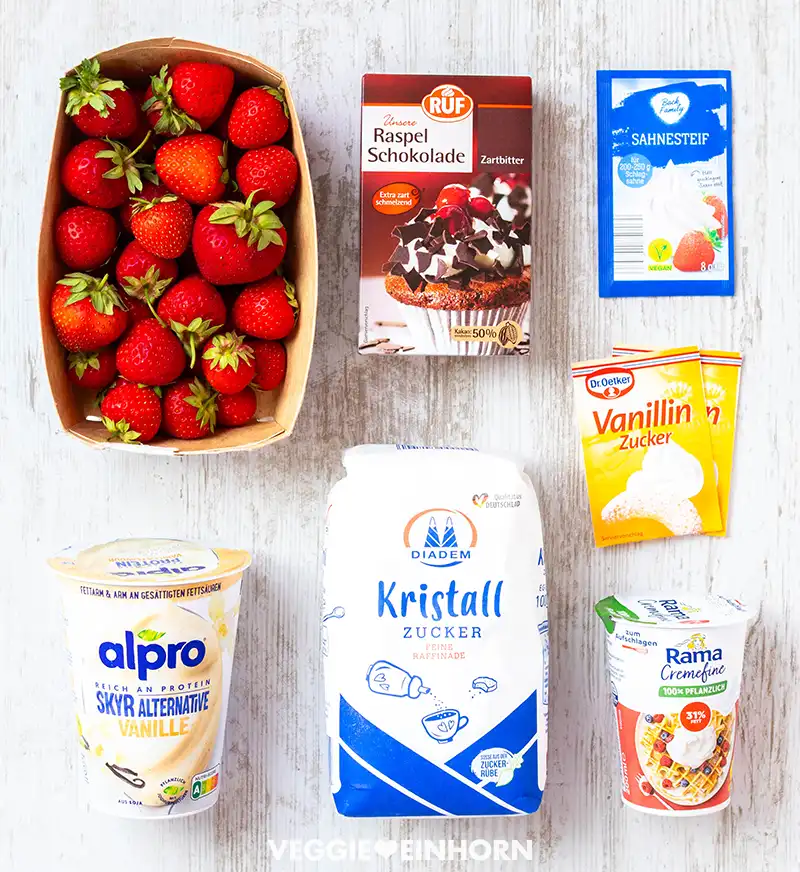 Zutaten auf einem Tisch arrangiert: Erdbeeren, vegane Raspelschokolade, Sahnesteif, Vanillinzucker,  veganer Vanille-Skyr, Zucker und vegane Schlagsahne.