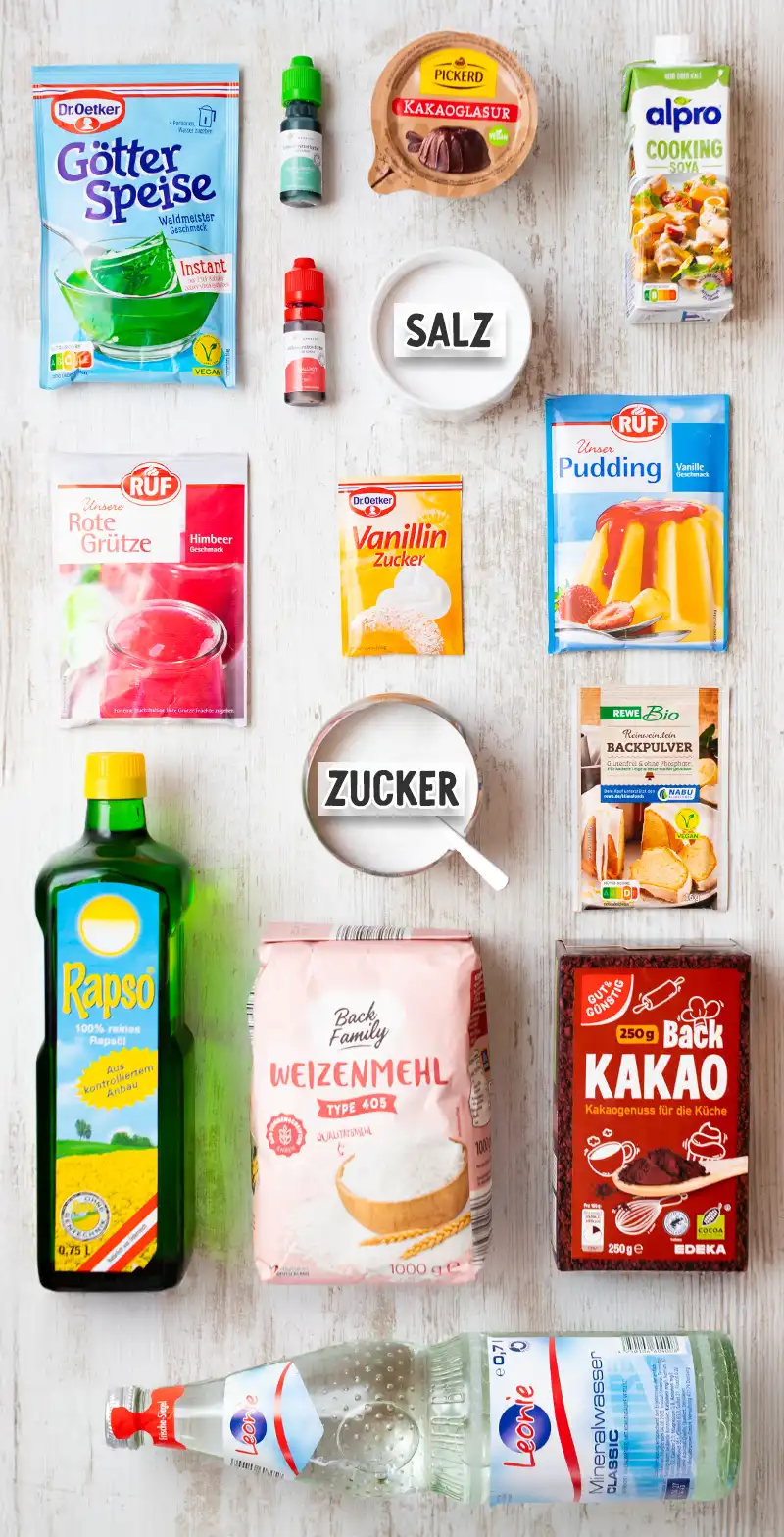 Zutaten für einen veganen Papageienkuchen auf einem hellen Holztisch arrangiert: Götterspeise Waldmeister, rote Grütze, Lebensmittelfarben in Grün und Rot, Kakaoglasur, Soja-Cuisine, Vanillinzucker, Vanillepuddingpulver, Backpulver, Salz, Zucker, Rapsöl, Weizenmehl, Backkakao und Mineralwasser.