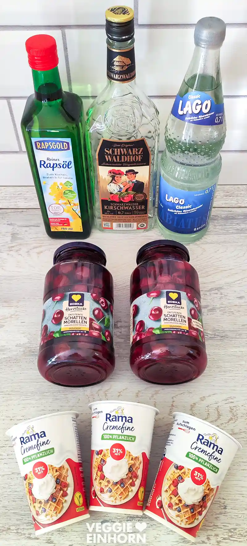 Zutaten für eine vegane Schwarzwälder Kirschtorte vom Blech. Zu sehen sind eine Flasche Rapsöl, eine Flasche Kirschwasser, eine Flasche Sprudelwasser, zwei große Gläser mit Schattenmorellen und drei Becher pflanzliche Rama Cremefine zum Aufschlagen.