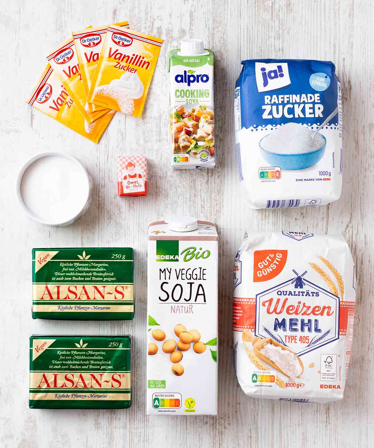 Zutaten für veganen Hefe-Streuselkuchen, darunter Mehl, Zucker, Vanillinzucker, frische Hefe, vegane Butter, Sojamilch, Soja-Kochsahne und Salz auf einem hellen Holztisch.